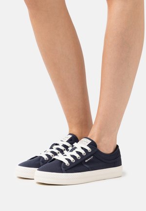Jambes portant des baskets basses GANT bleu marine avec des lacets blancs et des semelles en caoutchouc, debout sur un fond blanc.