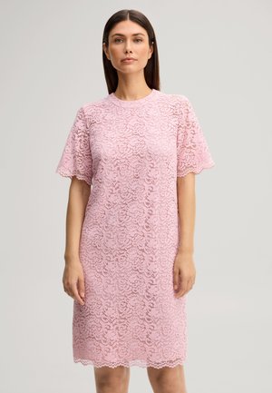 Donna in piedi che indossa un vestito di pizzo rosa a maniche corte e lunghezza al ginocchio, su uno sfondo grigio chiaro uniforme.