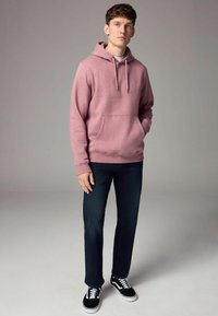 Sudadera rosa con bolsillo frontal y cordones grises, combinada con jeans oscuros y zapatillas negras, de pie frente a un fondo neutro.