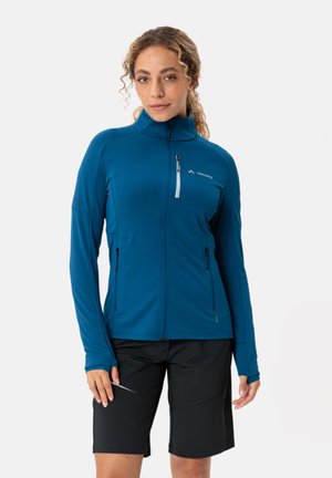 Blaue Zip-up Jacke mit hohem Kragen, zwei Seitentaschen und einer Brusttasche. Kombiniert mit schwarzen Shorts. Leichtes, dehnbares Material.
