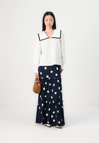 Hvit bluse med stor marineblå krage, kombinert med et marineblått skjørt med polka prikker og lagdelte volanger. Modellen holder en brun håndveske.