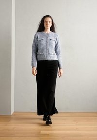 Un cardigan en tricot gris avec un col rond, deux poches avant et une fermeture à boutons, associé à un pantalon noir à jambes larges et des chaussures noires, le tout sur un fond uni.