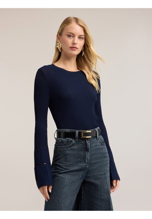 Mujer rubia con un suéter de canalé azul marino con mangas acampanadas, cinturón negro, vaqueros oscuros de talle alto y pendientes dorados en forma de hoja sobre un fondo liso.