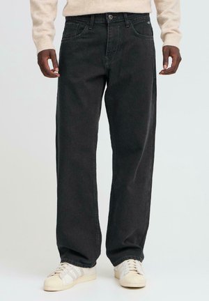 Homme portant un jean noir ample et des baskets blanches, debout avec les mains détendues le long du corps devant un fond uni.