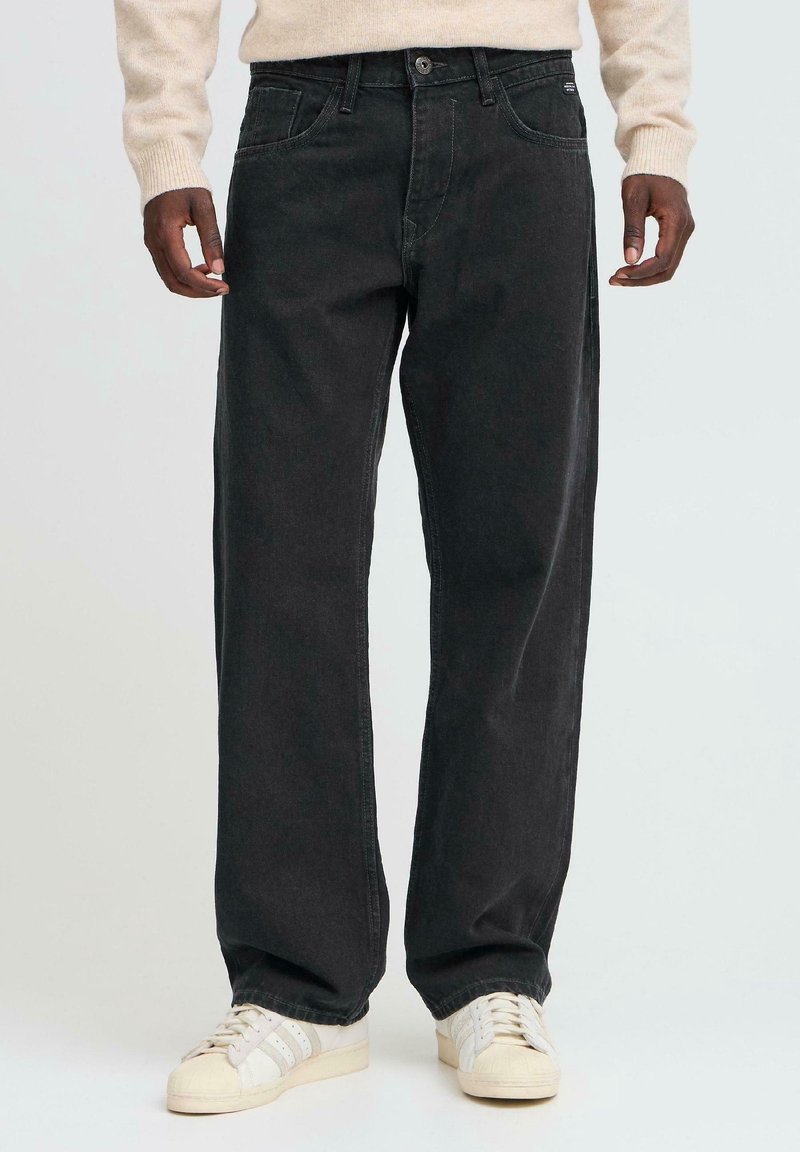 Homme portant un jean noir ample et des baskets blanches, debout avec les mains détendues le long du corps devant un fond uni.