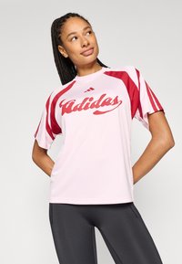 Rózsaszín sportos póló piros részletekkel, rövid ujjakkal és nagy "Adidas" logóval az elején. Simább, könnyű anyagból készült.