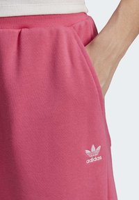 Pantalons de survêtement en coton rose avec une taille élastique, des poches latérales et un logo Adidas blanc sur la jambe gauche. Texture, coupe décontractée.