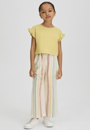 Ung pige står iført en gul bluse med flæseærmer, flerfarvede bukser med lodrette striber og hvide sneakers på en ensfarvet baggrund.
