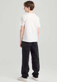 Valkoinen puuvillapaidan lyhythihainen t-paita, yhdistettynä mustiin löysiin farkkuihin ja valkoisiin lenkkareihin. Malli on kääntynyt pois päin, näyttäen taaksepäin.