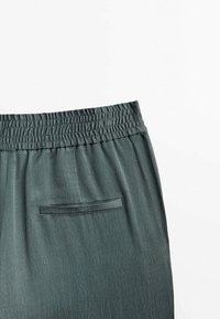 Partie de pantalon avec ceinture élastique vert foncé et une seule poche passepoilée horizontale sur tissu lisse sur fond blanc.