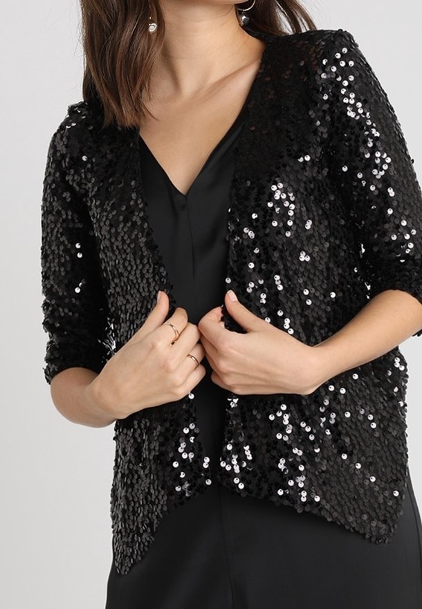 Femme portant une veste noire à sequins sur une tenue noire à col en V, tenant les bords de la veste avec les deux mains près de la poitrine.