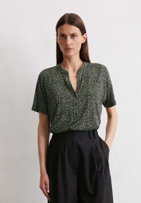 Donkergroene blouse met korte mouwen, voorzien van een textuur met een abstract wit patroon en een V-halslijn, gecombineerd met hooggetailleerde zwarte broek.
