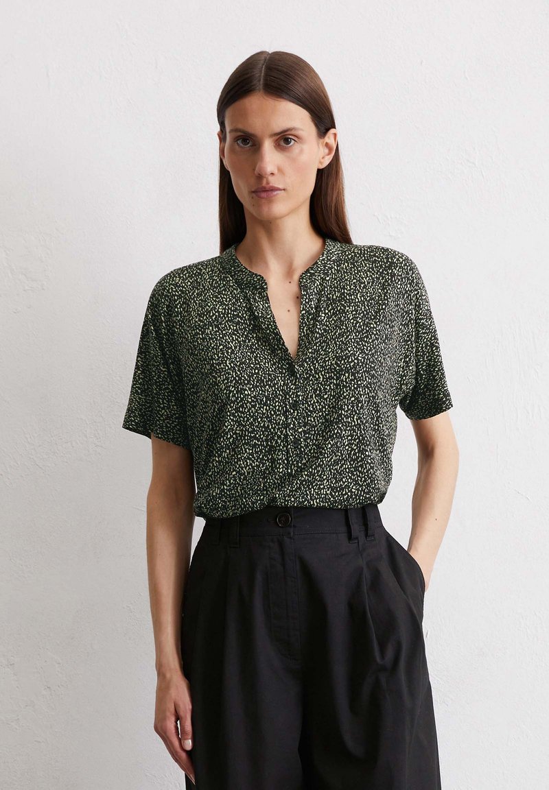Donkergroene blouse met korte mouwen, voorzien van een textuur met een abstract wit patroon en een V-halslijn, gecombineerd met hooggetailleerde zwarte broek.