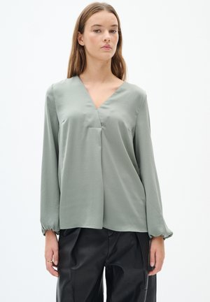 Blusa verde leggera con scollatura a V, maniche lunghe e vestibilità rilassata. Il materiale ha una texture liscia, con una leggera lucentezza.