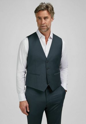 Mand med gråt hår og skæg iført en mørkegrå vest og bukser over en hvid skjorte, stående med den ene hånd i lommen.