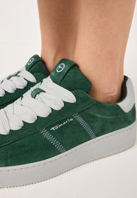 Sneaker verde in camoscio con lacci bianchi, caratterizzata da una suola in gomma grigia e logo sul lato. Superficie testurizzata con dettagli di cucitura a contrasto.