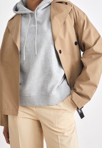 Cappotto trench beige sopra un felpa grigia chiara con lacci bianchi, abbinato a pantaloni color crema, con dettagli hardware visibili.