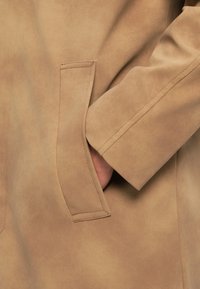 Beige faux suede coat with a smooth texture, featuring a side pocket and a wide cuffed sleeve, showcasing precise stitching details.