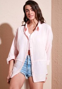 Light pink button-up shirt with a relaxed fit, made of textured fabric, featuring long sleeves and front pockets, layered over a white crop top.