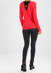 Fuksjowy sweter z dzianiny z głębokim dekoltem na plecach, odsłaniający czarną warstwę podstawową, zestawiony z czarnymi rurkowymi dżinsami i botkami na obcasie.