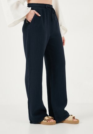 Persona che indossa pantaloni neri a vita alta, ampi e testurizzati, con cintura elastica e coulisse, abbinati a sandali beige con plateau.