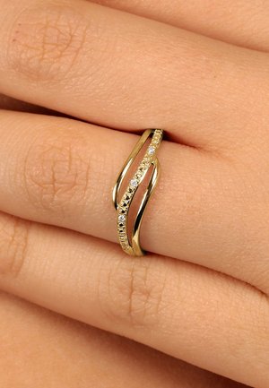 Goldring mit verwobenen Bändern, die kleine Diamanten enthalten, getragen an einem Finger vor einem neutralen Hauttön hintergrund.