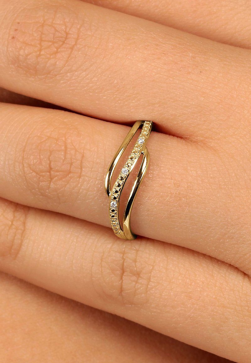Goldring mit verwobenen Bändern, die kleine Diamanten enthalten, getragen an einem Finger vor einem neutralen Hauttön hintergrund.