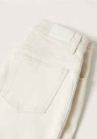 Jean en denim blanc cassé plié avec une poche arrière et une étiquette rectangulaire blanche sur la ceinture, sur un fond uni.