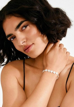 Zilveren armband met cirkelvormige schakels met gestructureerde accenten, gedragen om een blote pols tegen een neutrale achtergrond.