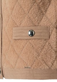 Close-up van beige textuur gebreide stof met een ruitpatroon, een drukknoop op een horizontale band en een klein metalen label dicht bij de onderkant.