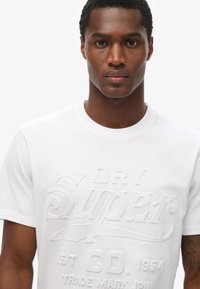 Hvid bomulds-T-shirt med præget tekstdesign, rund hals, korte ærmer og en glat tekstur. Ingen synlige mønstre eller accenter.