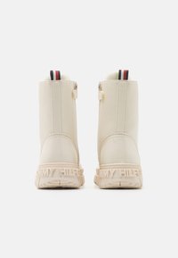 Beige enkellaarzen met een glanzende leren afwerking, een gestructureerde zool en een logo-detail op de hak. Voorzien van een contrasterende gestreepte trekband.