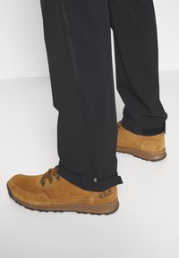 Bruine suède wandelboots met zwarte veters, gecombineerd met zwarte, taps toelopende outdoorbroeken met een zijrits aan de zoom.
