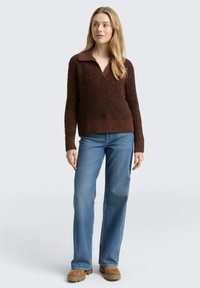 Bruine gebreide trui met een V-hals en kraag, gecombineerd met lichtblauwe uitlopende spijkerbroek en bruine klompen. Zachte stof, geribbelde zoom en manchetten.