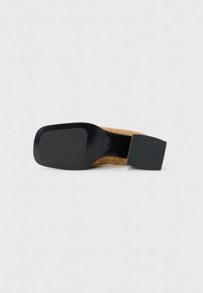 Chaussure à talon haut en suède beige avec un talon carré en cuir verni noir et une semelle extérieure lisse noire. Présente un design minimaliste.