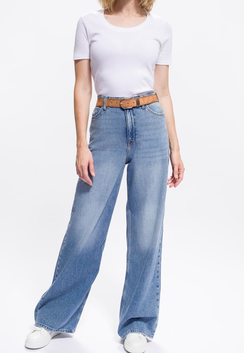 Frau trägt ein weißes enges Kurzarmtop, hochgeschnittene, weit geschnittene blaue Jeans mit brauem Gürtel und weiße Sneakers vor weißem Hintergrund.