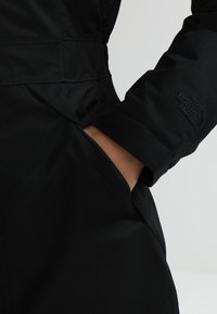 Sort isoleret jakke med en glat, mat finish; har en lynlås lomme og prægede logo på ærmet. Nærbillede af hånd i lommen.