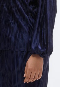 Μπλε σκούρο πλισέ τοπ με απαλή, γυαλιστερή υφή. Διαθέτει μανσέτα με πιέτες και χαλαρή σιλουέτα, αναδεικνύοντας ρέουσες γραμμές και πτυχώσεις.