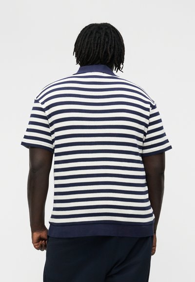 Bărbat cu dreadlocks scurte, purtând un tricou polo cu mânecă scurtă în dungi orizontale albastre și albe, cu fața întoarsă, pe un fundal simplu.