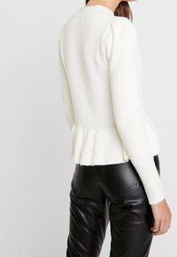 Valkoinen ribbineulottu neulepusero, jossa on puffihihat ja pepumhelma, yhdistettynä mustiin nahkahousuihin, jotka näyttävät sileältä ja istuvilta.