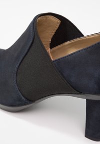 Gros plan d'une chaussure en daim bleu marine avec un panneau latéral élastique noir et un petit talon bloc sur fond blanc.