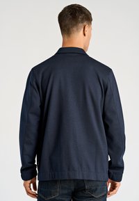 Granatowa kurtka z miękkiego materiału, z kołnierzem, długimi rękawami i delikatnymi detalami przeszyć. Widok od tyłu.