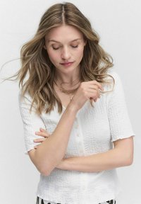Vit texturerad blus med korta ärmar, med en knapplist fram. Modellen poserar med armarna i kors, hår som faller i mjuka vågor.