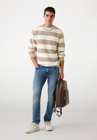 Pull en tricot rayé beige et crème, jeans bleus et baskets blanches. Sac à dos en cuir marron foncé avec une poche avant.