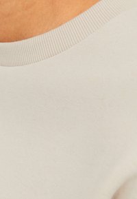 Lichtbeige katoenen top met een gladde textuur en een ribbelachtige ronde hals. De stof lijkt zacht en heeft een naadloze constructie.