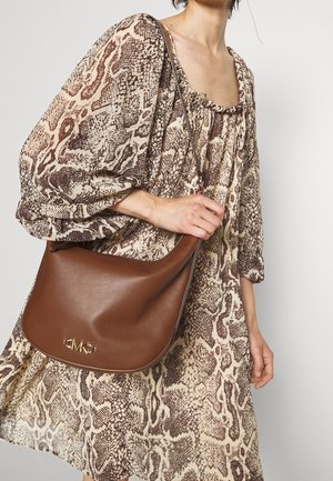 Sac à bandoulière en cuir marron avec une texture lisse et un détail logo doré, porté avec une robe à motifs dans des tons neutres.