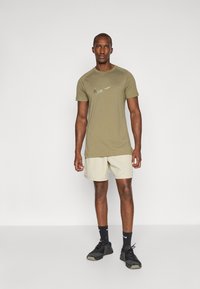 Olive green Nike t-shirt with a reflective logo, paired with beige shorts and black athletic shoes, standing on a neutral background.
