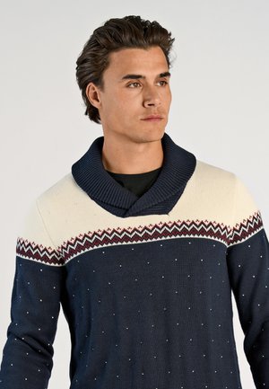 Stickad tröja i marinblått med krämfärgade detaljer, med sjalkrage, geometriska mönster och små vita prickar över hela.