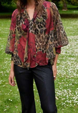 Blusa ligera y floral en rojo y verde con detalles de estampado animal. Presenta un diseño fluido y un cuello con lazo. Combinada con pantalones negros.