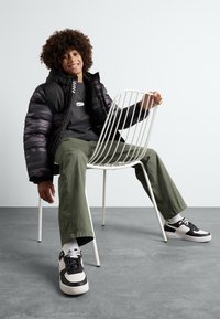 Chaqueta de plumas negra y gris, sudadera gris, pantalones verdes y zapatillas negras/blancas. Sentado en una silla de alambre blanca sobre un suelo gris.